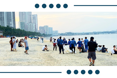 People enjoy a sandy beach along a calm bay, with groups and individuals near the water and a city skyline on the horizon.