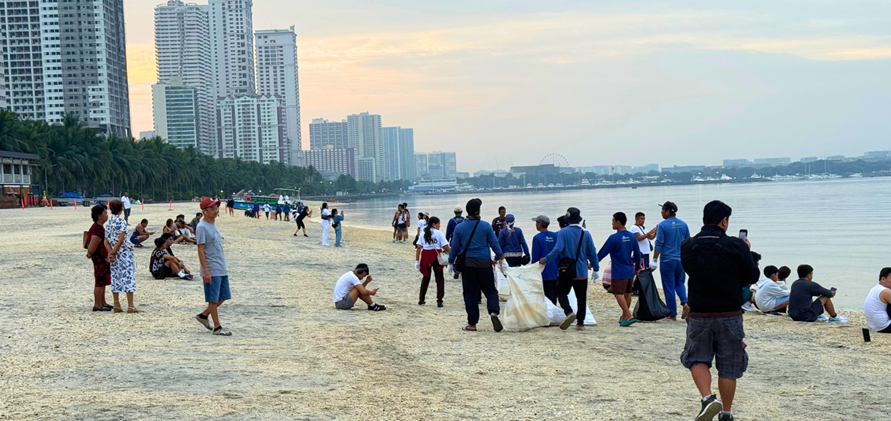 Crowded urban beach at sunset with high-rise buildings, palm trees, and people walking along the shore. Group of volunteers in blue shirts collecting litter on a sandy beach while others relax nearby. People enjoying a seaside cityscape: skyline, a distant ferris wheel, and calm water. Man takes a photo of a beach cleanup with a smartphone while others sit and stand along the shore. Wide beach scene showing volunteers picking up trash, pedestrians, and tall apartment blocks in the background.