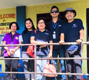 PTC FCR and RSE Foundation-2 | PTC Five adults pose behind a railing outside a yellow building, wearing navy shirts with lanyards and badges, smiling for a group photo.