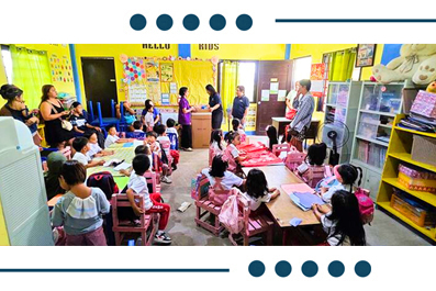 Kindergarten classroom with children seated at small desks while teachers lead a lesson at the front of the room.