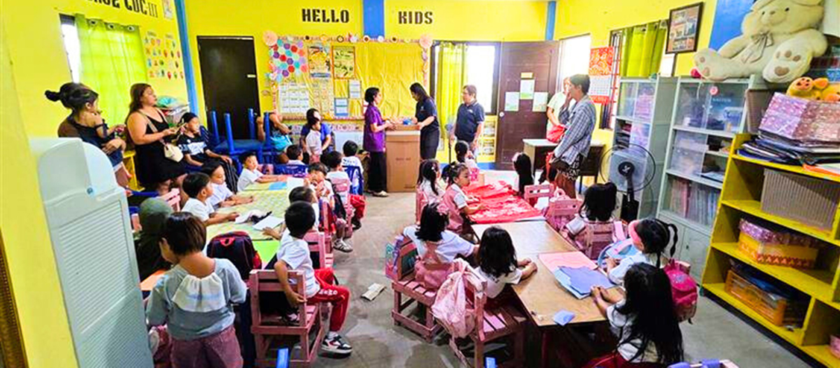 PTC FCR and RSE Foundation | PTC Kindergarten classroom with many children seated at small desks while adults stand at the front supervising; bright yellow walls display 'HELLO KIDS'.