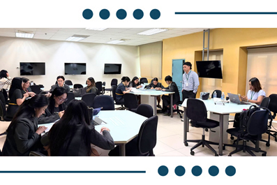Busy classroom with students seated around tables, some using laptops, while an instructor stands at the front of the room.