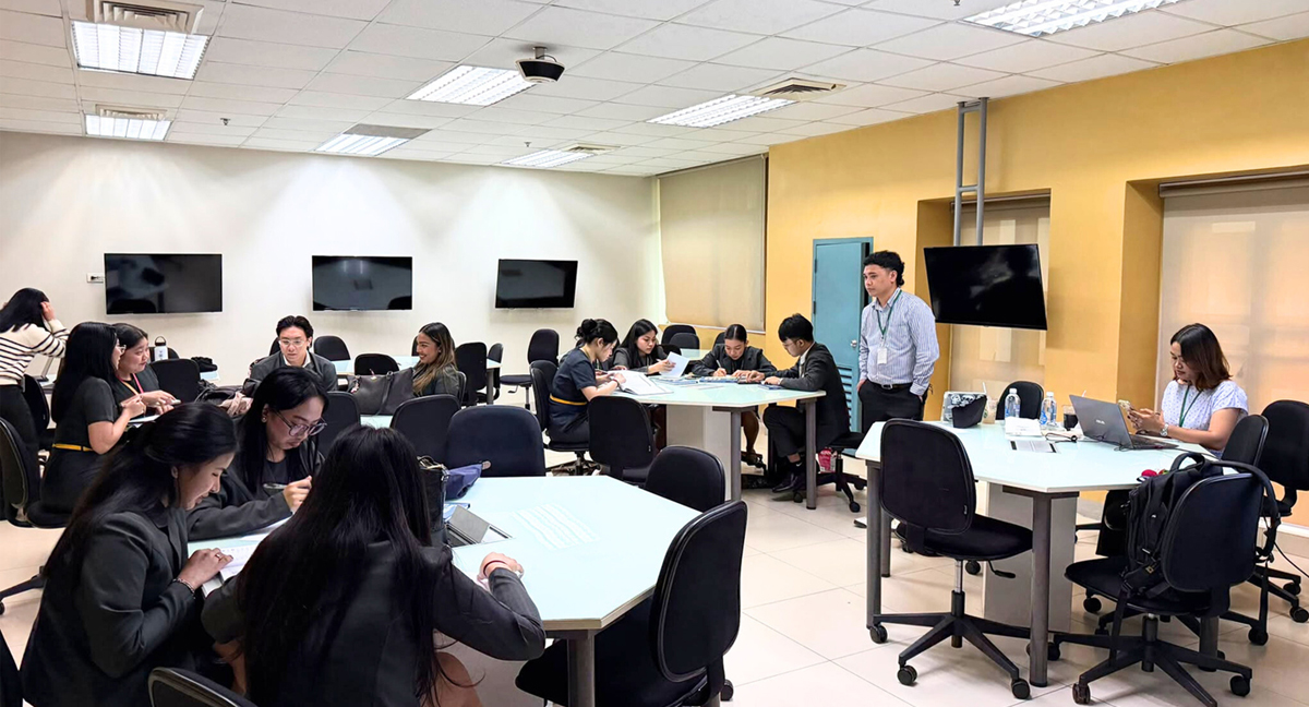 Group of professionals seated around tables in a bright training room, a man stands at the front as instructor.
