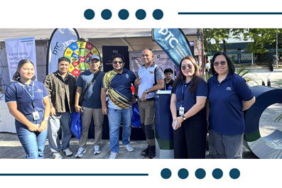 Group of people posing outdoors at a promotional event with a prize wheel and banners in the background.