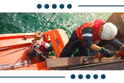 Crew members in hard hats and life jackets prepare a rescue raft on a bright orange boat above turquoise water.