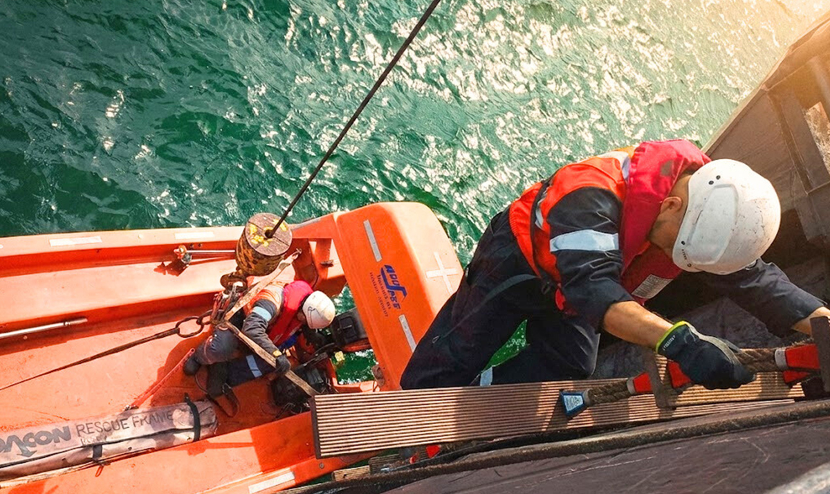 Two rescue workers in orange life jackets and helmets secure a metal beam on an orange rescue boat above green water, rope and pulleys nearby.