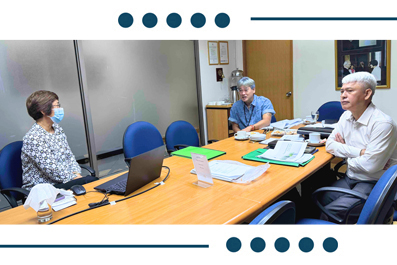 Four people sit around a wooden conference table in a meeting; one person wears a face mask and a laptop is open on the table.