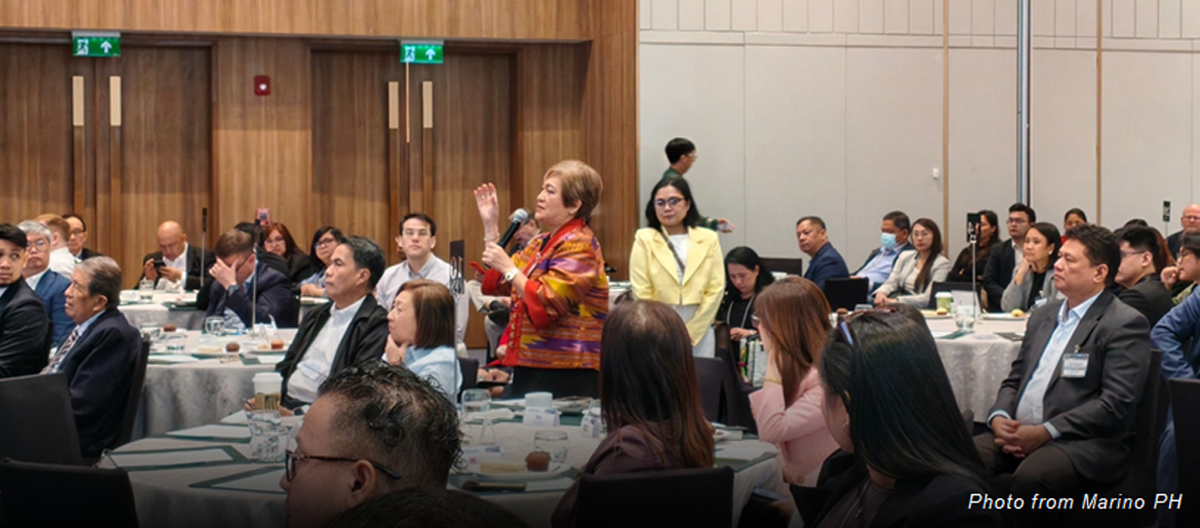 Woman standing with a microphone asking a question during a formal conference, surrounded by seated attendees at round tables in business attire.
