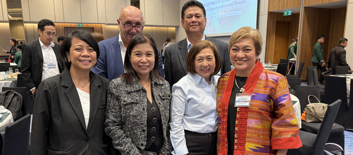 Six diverse professionals in business attire smile for a group photo at a conference, indoors with tables and a presentation screen in the background.