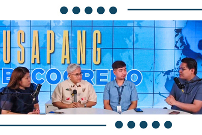 Panel discussion with four speakers seated at a table on a blue stage backdrop, each with a microphone and lanyards.