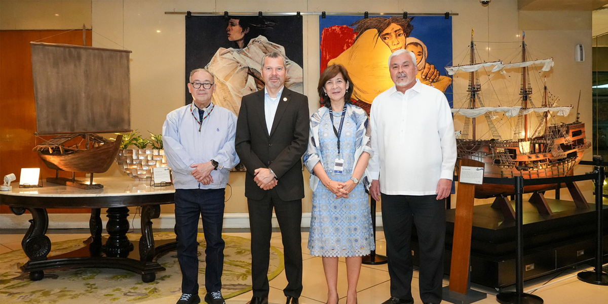 PTC hosts IMO Secretary-General | PTC Four adults stand together in a gallery, posing in front of large colorful paintings and a model ship display on both sides.