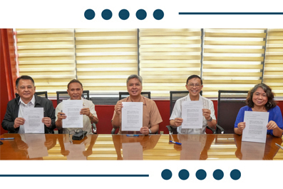Five people sit at a conference table holding up signed documents toward the camera, suggesting a signing ceremony.