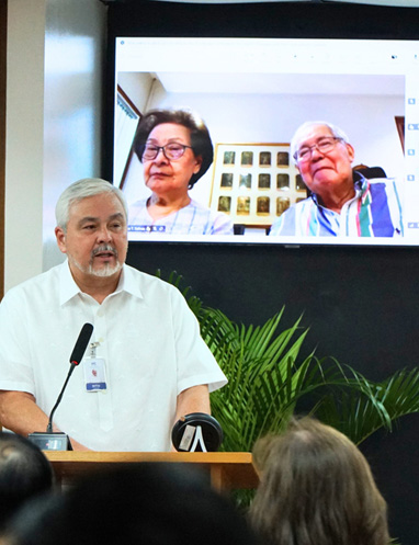 PTC marks 47 years-5 | PTC Man in a white shirt speaking at a podium with a microphone, addressing an audience; two older people appear on a large screen behind him during a video call