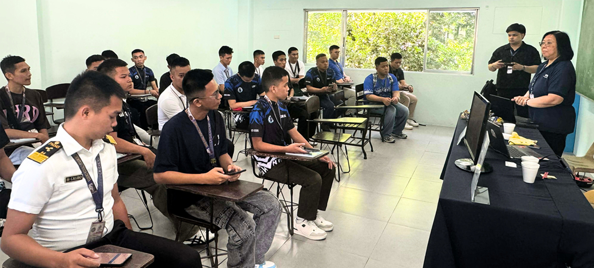 Classroom training session with students wearing lanyards facing two instructors at a front table with laptops and cups on the desk.