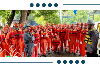 Group of workers in orange coveralls and hard hats listening to a supervisor in gray overalls during an outdoor safety briefing at a worksite.