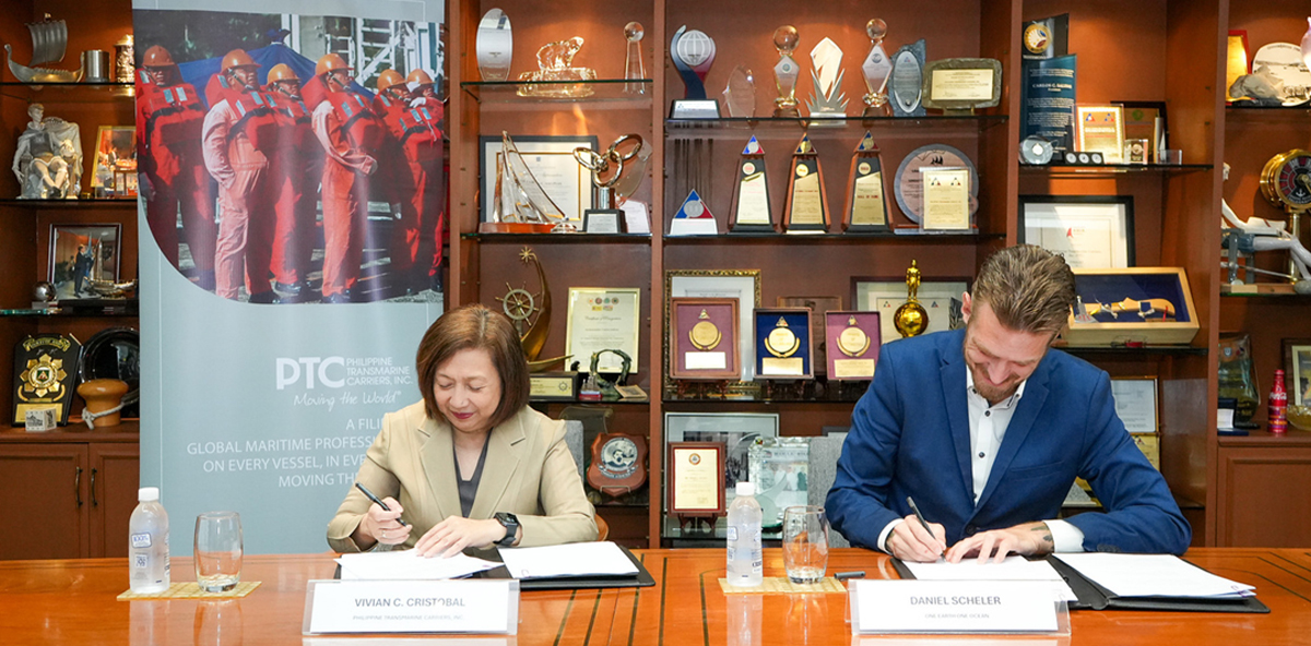 Two professionals sign documents at a formal signing ceremony, with a trophy-filled display cabinet behind them. The nameplates read Vivian C. Cristobal and Daniel Scheler.
