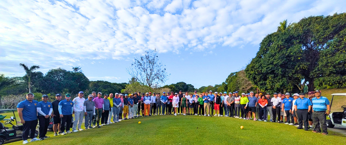 18th FAME Golf Invitational-2 | PTC Large group of people standing in a line on a sunny golf course, posing for a group photo with trees and blue sky in the background.