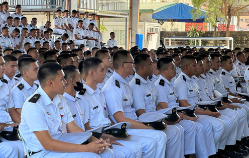 Midway Colleges-3 | PTC Group of military cadets in white uniforms seated in bleachers at an outdoor ceremony, hats on laps, with a blue canopy in the background.