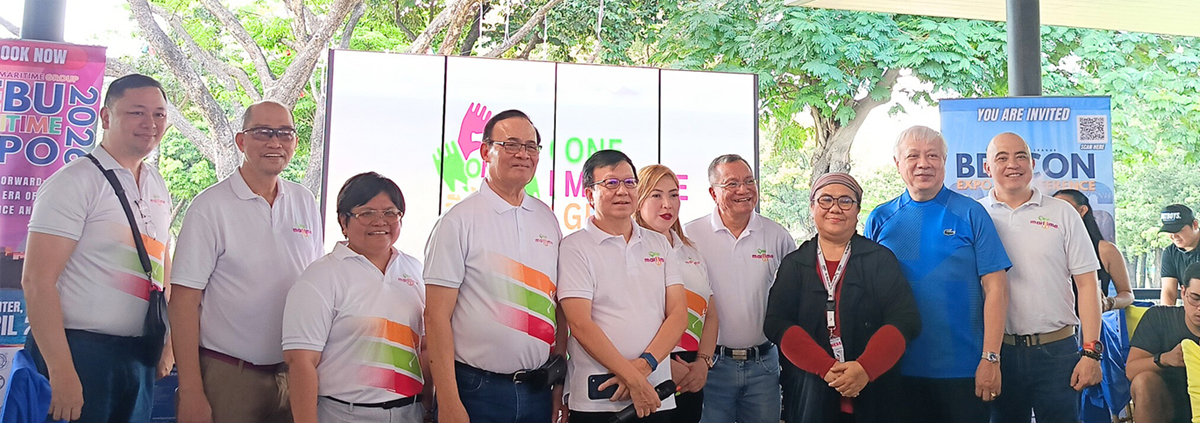 Group of nine adults posing for a photo at an outdoor expo, wearing white polo shirts with colorful diagonal stripes, banners and a screen behind them.