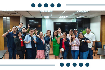 Group of about 20 colleagues posing for a photo in a conference room, some giving thumbs up, in colorful business casual attire.