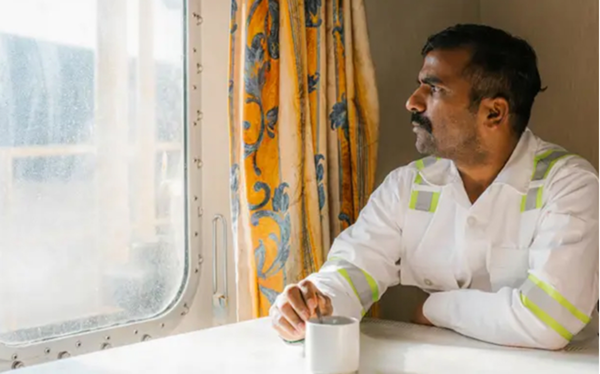 Man in a white uniform with reflective bands sits at a table by a train window, holding a mug and gazing outside.