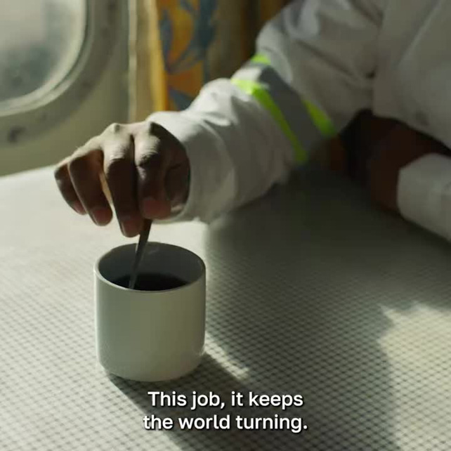 Person in a white uniform stirs a drink in a white mug on a metal surface; caption reads, 'This job, it keeps the world turning.'