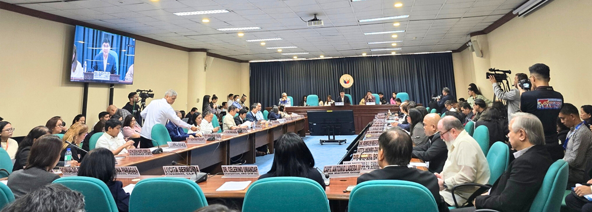 Large conference room with officials seated around a U-shaped table, press filming in the back and a screen showing a speaker on the left edge of the frame.