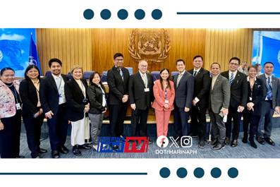 Group of about fifteen professionals in business attire posing for a formal group photo in front of a government emblem in a conference room.