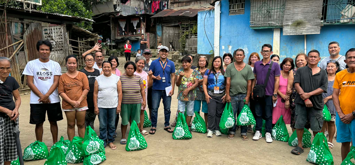 Beyond borders-2 | PTC A diverse group of adults standing in a line on a dirt street, each with a green gift bag, during a community aid event.