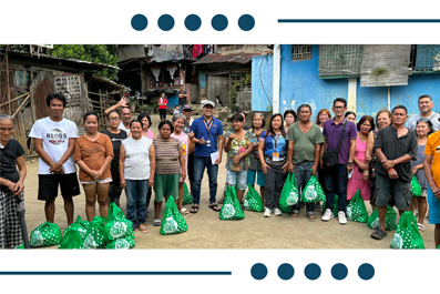 Group of about 20 people standing on a village street with green gift bags, posing for a community aid event.