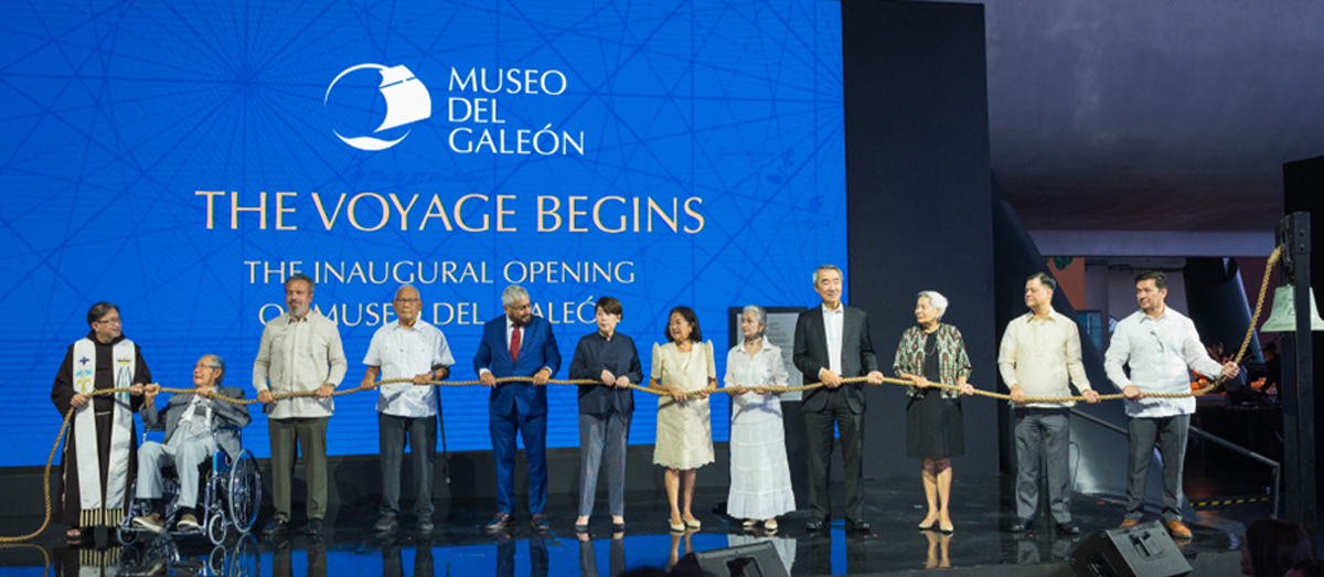Line of diverse officials and guests on stage holding a ceremonial rope at the Museo del Galeón opening event, with a blue backdrop reading 'The Voyage Begins'.