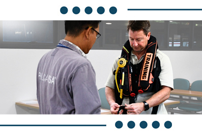 Instructor helping a trainee fasten a safety harness with a label reading 'TRAINING' in a classroom setting.