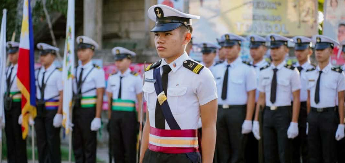 Uniformed cadet in white shirt and peaked cap leads a marching squad during a parade, with fellow officers behind him.