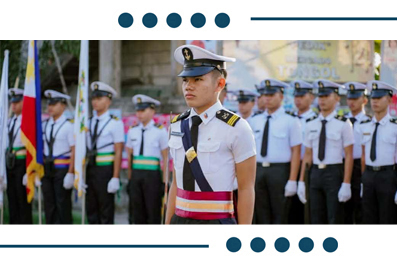 Young cadet in a white naval dress uniform leads a line of uniformed cadets at a ceremony, with flags in the background.