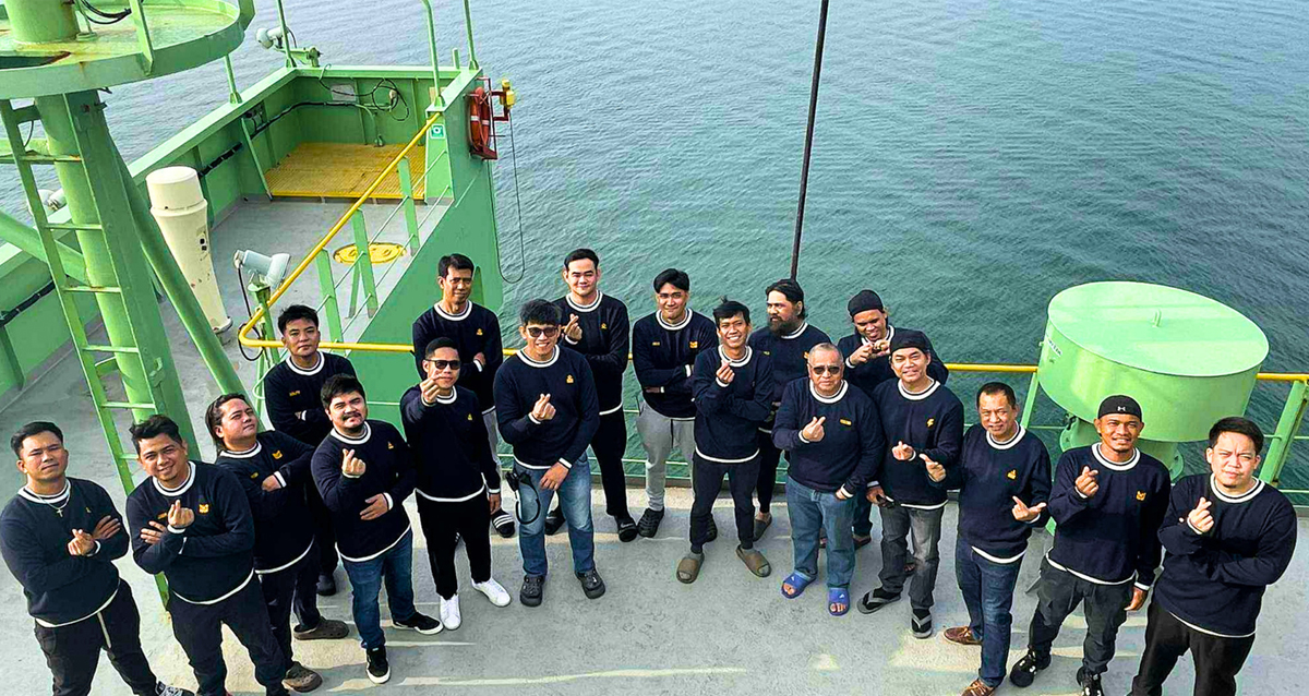 the Filipino maritime family-2 | PTC Group of ship crew members in matching dark sweaters pose on a green deck above blue water, making finger-heart gestures at the camera.