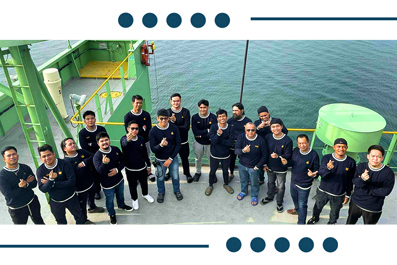 Group of about 20 people standing on a ship deck, giving thumbs up, with water in the background.
