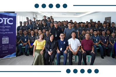 Group portrait of around 40 people seated and standing in a conference room, with a blue OTC banner on the left.