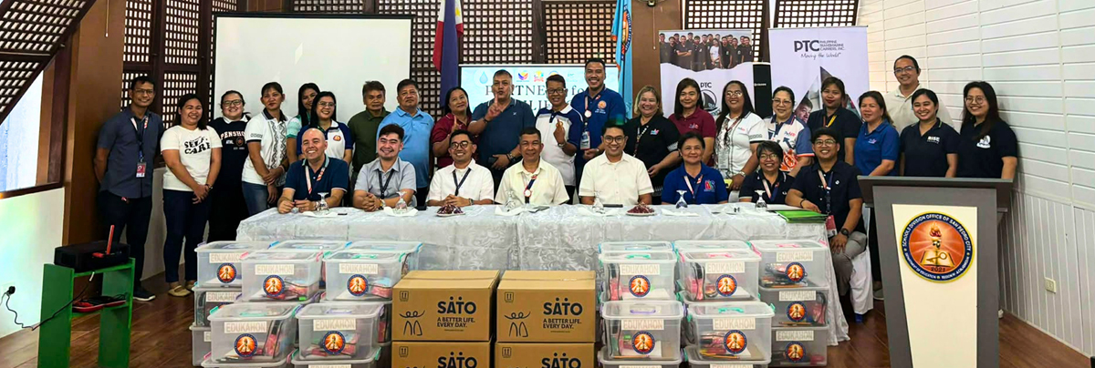 Group of about 30 people at a community charity event posing for a photo behind a long table with stacked donation boxes and supplies; banners and a podium are visible in a bright room.