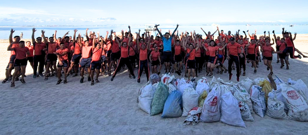 One Earth One Ocean and PTC advance marine-2 | PTC Large group of volunteers in red shirts on a sandy beach, posing with filled bags of collected trash in front of the shoreline.