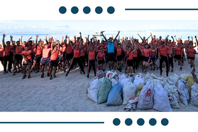 Group of volunteers on a beach posing with filled trash bags after a cleanup, ocean in the background.