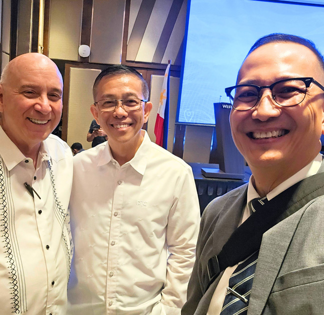 Three men in a hallway posing for a selfie at a conference, smiling with a stage and Philippine flag in the background.