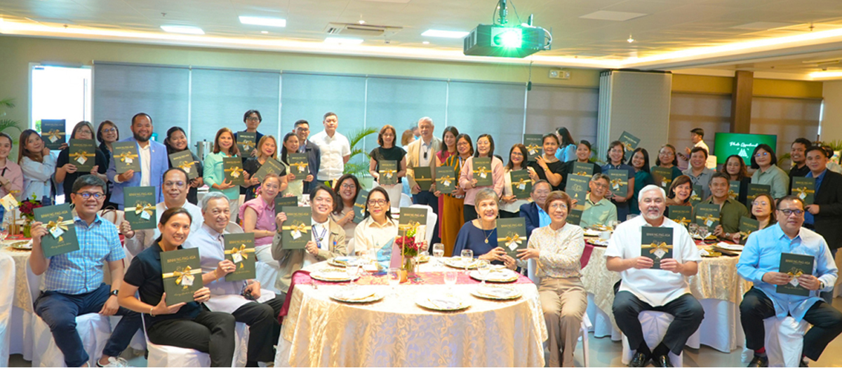 Large group of people at a banquet posing for a photo, each holding a green booklet with a gold bow, around round tables set for a meal in a conference room.