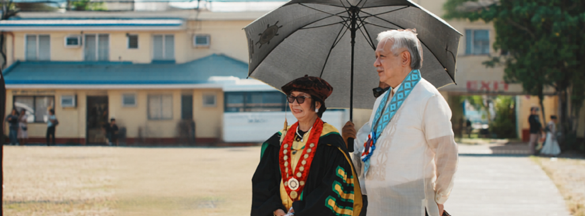 challenges graduates to stay grounded-2 | PTC Two seniors in ceremonial dress walk outdoors; woman wears a red-sashed regalia with medallion, man holds a gray umbrella.
