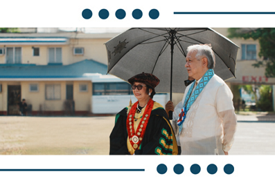 Two dignitaries in ceremonial robes outdoors: a man in white with a black umbrella stands beside a woman wearing a colorful ceremonial gown and sash, with a light building in the background.