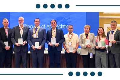 Eight professionals in business attire pose with certificates and medals on a stage during an awards ceremony. The backdrop shows a conference setting.