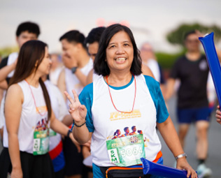 17th FAME Fun Run 2026-7 | PTC Smiling woman in a white t-shirt with a race bib, blue cardigan, and peace sign at a running event; participants in the background.