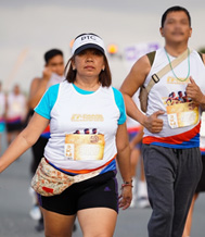 17th FAME Fun Run 2026-8 | PTC Female runner in a blue shirt leads a road race, wearing a white bib and black shorts, with a male runner in a white shirt and blue shorts beside her; both carry race bibs in a crowded outdoor finish or start area.