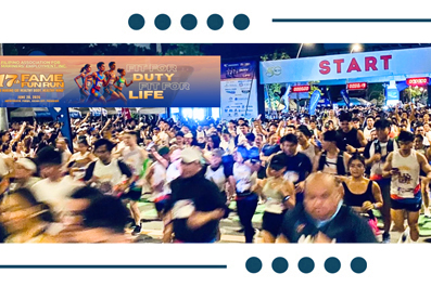 Crowd of runners at the race start, with a prominent START banner overhead and event banners in the background.