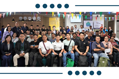 Group of people posing for a conference group photo, many wearing lanyards, with colorful bunting overhead and backpacks at their feet.