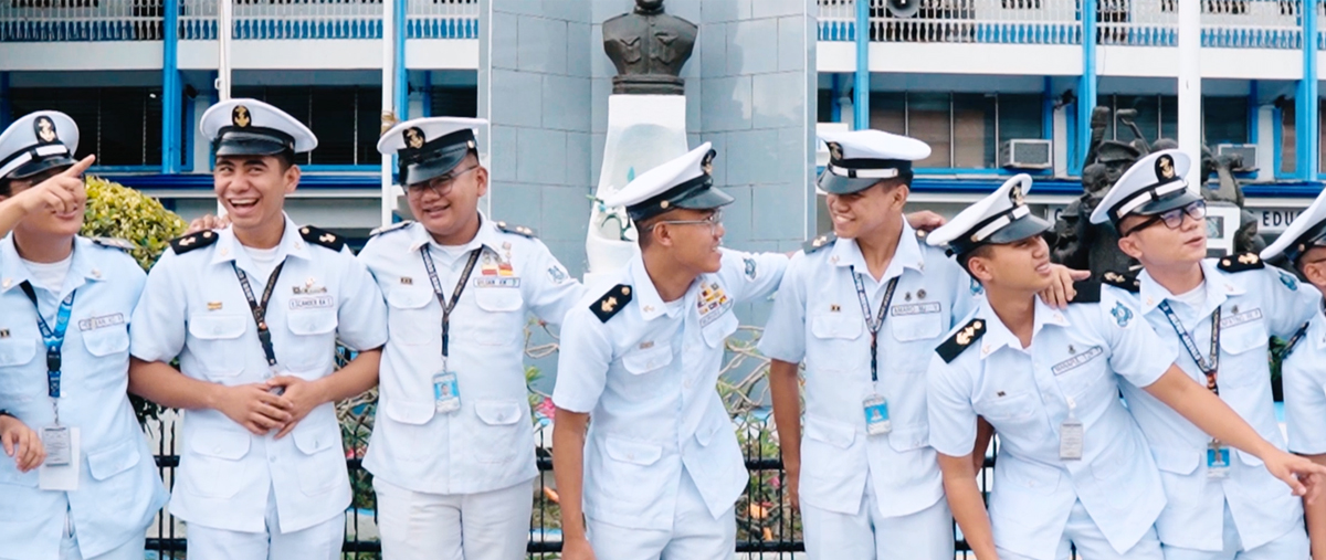 Group of uniformed sailors standing together outdoors, smiling and linking arms in light-blue dress uniforms.